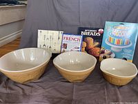 Three beige ceramic mixing bowls of graduated sizes with embossed exterior pattern, displayed with four cookbooks behind them on grey fabric background.