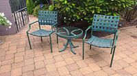 Full view of two green metal patio chairs and round glass-top table arranged outdoors on a paved surface with green foliage in the background.