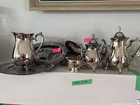 Full set of silver plated items displayed on a white surface including coffee pot, teapot, water jug, two trays, salad servers, and cream sugar set. Visible markings and condition noted.