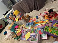 Overview of assorted baby and children's toys, puzzles, and plush soft toys arranged on floor with boxes and bags visible.