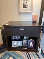 Full photo of the dark wood laminate bedside table with drawer and open lower shelf containing picture frames.