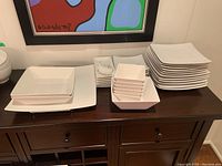 Stacked white square ceramic plates and bowls with matching white spoons on a dark wooden table, showing quantity and size variety.