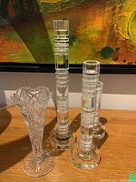 Three clear crystal candle holders with geometric, stepped designs and circular bases next to a tall clear crystal vase with intricate etched floral and geometric patterns and scalloped rim on a wooden surface