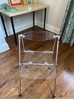 Unfolded chair showing transparent Lucite backrest and seat with chrome tubular legs and structure on hardwood floor.