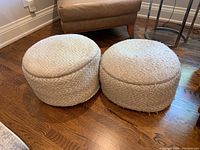 Two round fabric-covered footstools or poufs on hardwood floor near chairs and table, showing both top and side views with textured/woven designs.