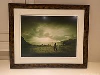Framed photographic print showing the full artwork with double matting and wooden frame, depicting Gaucho walking in landscape with mountains and cloudy sky.