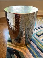 Full front-side view of the hammered metal cylindrical table on a colorful rug with visible hammered texture and reflective surface.