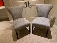 Front view of two matching upholstered chairs with ribbed grey fabric upholstery and dark wood legs on carpeted floor in front of double doors.