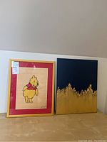 Both paintings shown side by side: framed Winnie the Pooh illustration in gold frame with pink mat; abstract navy and gold acrylic painting unframed.