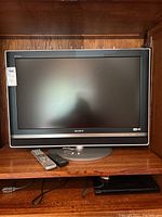 Full view of Sony Bravia TV on wooden shelf with two remotes in front and DVD player below