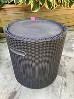 Closed resin weave round outdoor table with solid removable top on stone patio beside plants.