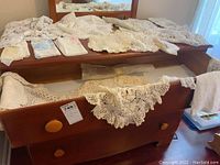 Wide view of dresser top and open drawer displaying numerous crocheted scarves, doilies, and handkerchiefs in cream and white shades.