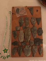 Wooden board with 23 various genuine stone arrowheads mounted plus a small loose plastic bag with two additional arrowheads beside a beaded necklace with a flower design