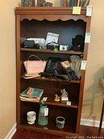 Front view of wooden bookcase with four shelves containing various items like cameras, purses, small figurines and miscellaneous objects; bookcase shows scratches and use.
