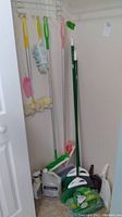 Cleaning tools including dusters with yellow, green, and pink handles, mop with green and white handle, brooms, dust pans, and bottles of cleaning liquids arranged in a corner of a room.