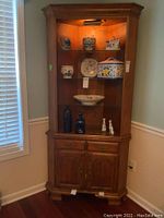 Full view of the Hale Maple corner cabinet fitted into the corner with light on, glass doors showing decorative ceramics on shelves, two drawers and double-door cabinet below.