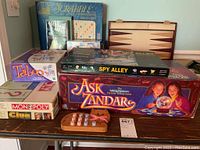 Overall lot of assorted classic board games stacked on table including Scrabble Deluxe, Backgammon board, Monopoly, Clue, Taboo, Spy Alley, Ask Zandar, and Master Mind with visible game boards and boxes.