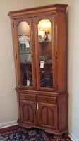 Front view of the maple corner cabinet showing two glass doors with visible shelves and contents inside. Lower drawer and cabinet doors also visible. Shows wood finish and general condition.