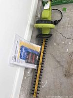 Photo of electric hedge trimmer lying on floor showing 22-inch blade and green body with black guard and power cord, original paperwork stored in clear plastic sleeve beside it.