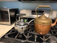 Photo showing two kettles on a gas stove; the subject kettle is the one on the left, a shiny chrome finished one; the other kettle on the right is solid copper with brass handle and spout, more patina.
