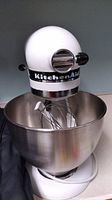 White KitchenAid Classic Plus stand mixer front view with stainless steel bowl and wire whisk attachment