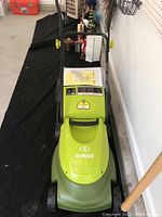 Front view of green Sunjoe electric lawnmower in garage on black carpet, showing brand logo, handle, and front wheels.