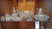 Full view of pressed glass and barware items including decanter, bowl, sugar server, pitcher, and shot glasses on a glass shelf