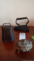 Photo showing cast iron sadiron, steel cow bell, and Rogers silver footed trinket box on wooden surface