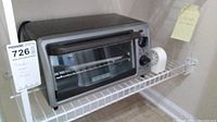 Front view of Black and Decker toaster oven on a wire shelf with kitchen items around it, including a white round vintage-style timer and small container.