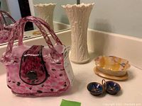 Photo shows the entire lot on a white countertop including a tall cream colored Lenox vase with floral embossing, pink and black blown glass purse, oval onyx box with lid, and small blue enamel trinket holder.