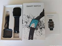 Smart watch, charging dock with cable, original packaging box, and quick guide manual displayed on white surface.