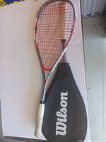 Wilson racquet laid flat showing head, strings, handle, and accompanying black protective cover with Wilson logo.