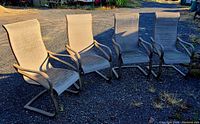 Four outdoor patio chairs arranged in a row on gravel ground, showing beige fabric and metal frames