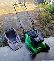 Full side view showing entire mower including grass catcher bag and wheels on concrete surface with gravel and grass in background.