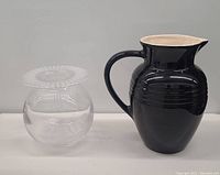 Black ceramic Le Creuset pitcher and clear glass vase side by side on a white surface against grey background.