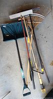 Photo of lawn and garden tools including rakes, a broom, a green plastic snow shovel, and a garden rake showing overall condition and variety