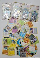 Photo of Pokémon card lot showing four small packets of holographic cards arranged in a line at the top, with a loose sprawling pile of mixed Pokémon cards (including some trainers and energy cards) below on a white surface.