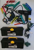 Top view of assorted hand tools including utility knives, scissors, screwdriver, tape measure, wire stripper, electric drill, heat gun, and two black plastic toolboxes with yellow locks and handles.