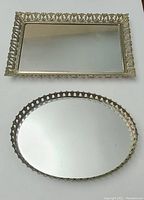 Top view of two vintage mirrored vanity trays, one rectangular and one oval, showing mirrored surfaces and decorative gold-tone frames.