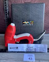 Red and white electric staple gun in front of its black Arrow Electro-Matic branded case with an instruction manual and a MaxSold bidding card in the foreground.