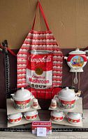Full view of apron with Campbell's Cream of Mushroom Soup design with pot and bowls on display stands, salt and pepper shakers, and soup can clock hung on wall.