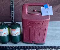 Photo showing two green Coleman propane fuel canisters and a maroon Coleman propane lantern case with handle and embossed logo.