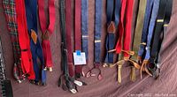 Overview of 13 men's dress suspenders in multiple colors and attachment styles laid out flat on a brown surface with a ruler for scale.