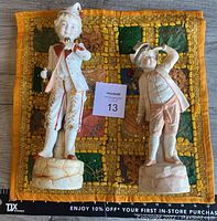 Two porcelain statues on a multicolored quilted cloth with the taller figure on the left and smaller figure on the right; taller statue appears reassembled.
