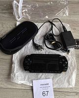 Photo showing the PSP console along with power supply, black zippered soft carry case, and clear plastic sleeve.