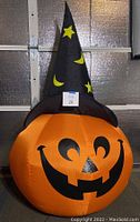 Front view of inflatable orange pumpkin with smiling jack-o-lantern face and black witch hat with yellow stars and crescents.