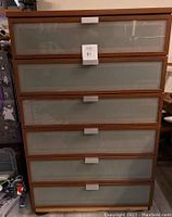 Front view of the IKEA dresser showing six drawers with glass fronts and wooden frames, with a white label on the top drawer center.