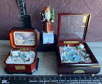 Two Thomas Kincaid Holiday Merriment music boxes and Santa figurine shown together on wood surface with measuring tape for scale.