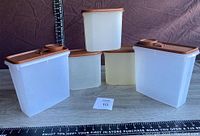 Five Tupperware containers with lids arranged on surface with measuring tape for scale. Three larger white translucent containers and two smaller cream translucent containers with brown lids.