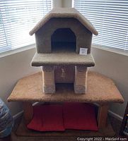 Front view of cat tree showing enclosed top house, carpeted platforms, and plist support posts with red cushions at the base.